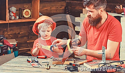 Fatherhood concept. Boy, child busy in protective helmet learning to use handsaw with dad. Father, parent with beard Stock Photo