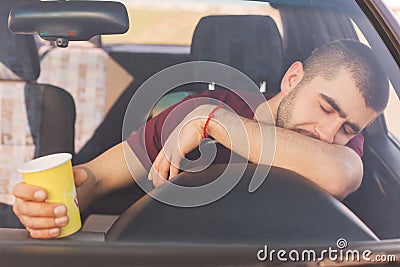 Fatigue overworked tired male driver has long trip in car, makes stop to have rest, has nap on helm, holds coffee in hands, looks Stock Photo