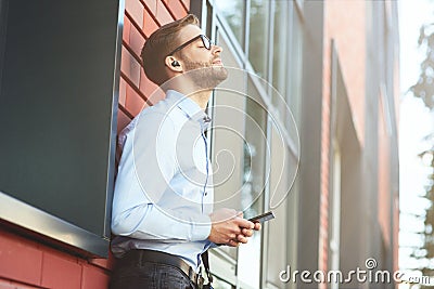 Favourite song. Side view of a young happy businessman listening to music with wireless earphones and mobile phone while Stock Photo