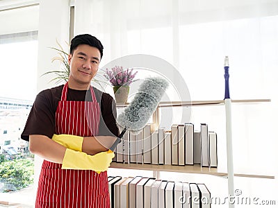 Feather duster in his hand while wearing yellow rubber gloves and sanitizer bottle spray for cleaning book shelf in living room at Stock Photo