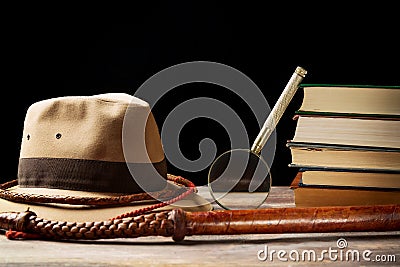 Fedora hat with bullwhip near magnifying glass and old books on black background. Adventure concept Stock Photo