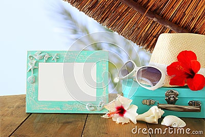 Fedora hat, sunglasses, tropical hibiscus flower next to blank frame over wooden table and beach landscape background Stock Photo