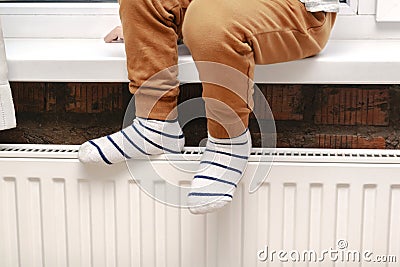 Feet legs of little boy kid in warm socks.Child sitting by window and warming up from heat radiator.Centralized heating Stock Photo