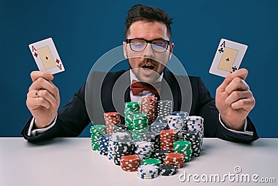 Fellow in glasses, black suit sitting at table with stacks of chips, holding two playing cards, posing on blue Stock Photo