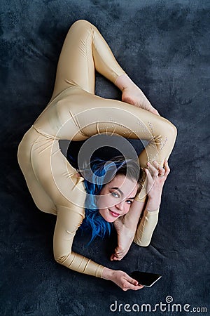 A female acrobat lies on a gray bedspread in a curved pose and uses a mobile. A woman contortionist resting on a bed in Stock Photo