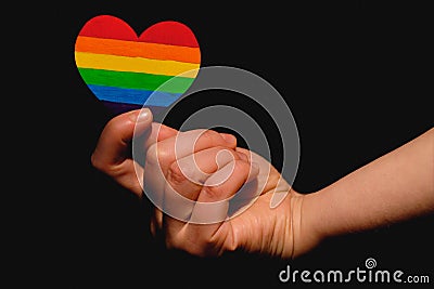 Female arms holding the heart coloured in LGBT pride colours on the dark background. Concept of the International Day Against Stock Photo
