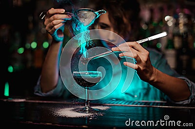 Female bartender pouring on the brown cocktail and on a flamed badian on tweezers a powdered sugar in the green light Stock Photo