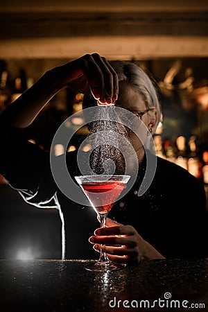 Female bartender sprinkling the orange peel into the cocktail glass filled with brown alcoholic drink Stock Photo