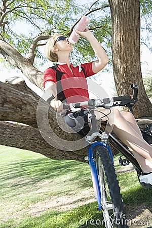 Female Bicyclist Sitting On Tree While Drinking Water Stock Photo