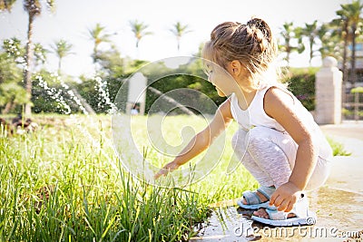 Female blonde cute child bent over the edge of a fountain in the park, touching the water jets with her hand - Concept: the Stock Photo