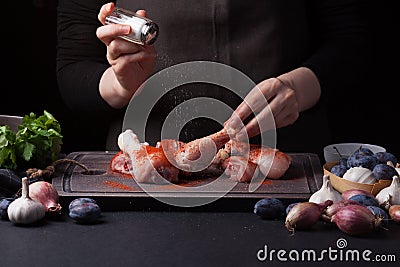 A female chef sprinkles fresh raw chicken drumsticks on a dark background with sea salt. Nearby lie the ingredients for Stock Photo