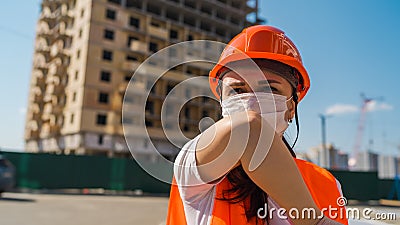 Female construction worker in overalls and medical mask coughing in elbow on background of house under construction Stock Photo