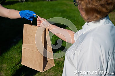 Female courier in gloves bringing food package to retired old woman. Woman handing a paper bag with groceries to a Stock Photo