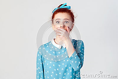 Female in despair and shock. Portrait of young desperate redhead girl in blue dress looking panic, covered her mouth by hand. Stock Photo