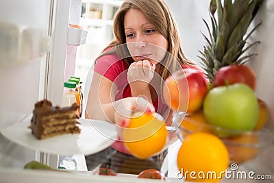 Female on diet in dilemma whether to eat chocolate cake or orange from fridge Stock Photo