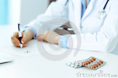 Female doctor filling up prescription form while sitting at the desk in hospital. Close-up of medical pills, lying at Stock Photo