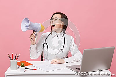 Female doctor sit at desk work on computer with medical document hold megaphone in hospital isolated on pastel pink Stock Photo