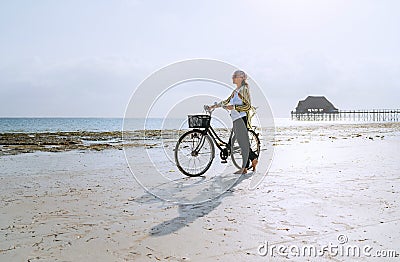 Female dressed light summer clothes have morning walk with old vintage bicycle with front basket on the lonely low tide ocean Stock Photo