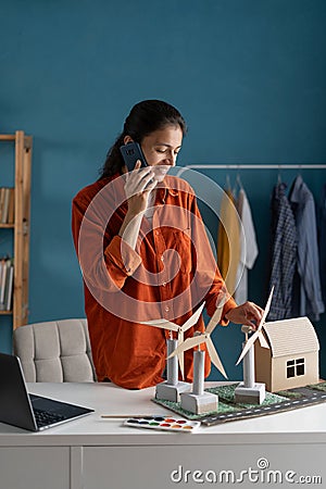 Female ecology engineer sitting at home and talking on the phone about project. On desk are windmills and house model Stock Photo