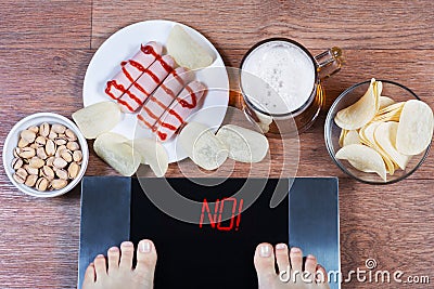 Female feet on digital scales with word no on screen. Beer and plates with junk food sausages, potato chips, pistachios. Stock Photo