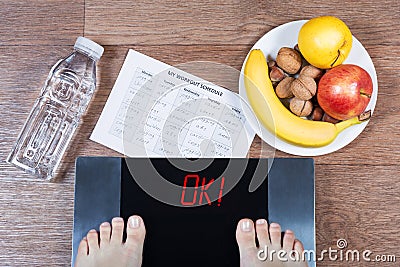 Female feet on digital scales with word ok surrounded by bottle of water, plate with healthy food and workout schedule paper. Stock Photo