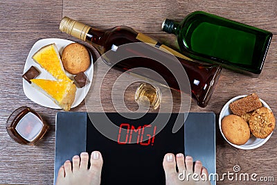 Female feet on digital scales with word omg on screen. Bottles and glasses of alcohol, plates with sweet food. Stock Photo