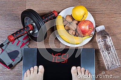 Female feet on digital scales with word yes surrounded by sport accessories AB roller wheel, fitness gloves, healthy food. Stock Photo