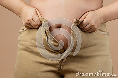 Female flabby big tummy preventing her from putting jeans on on beige background. Visceral fat. Body positive. Sudden Stock Photo