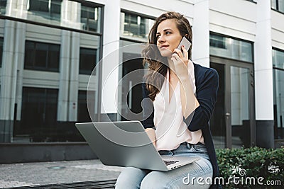 Female freelance working with laptop on street in business district and discusses work by phone with customer Stock Photo