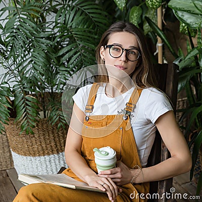 Female gardener in glasses resting, sitting on wooden chair in home greenhouse, hold collapsible silicone eco muge coffee/tea mug Stock Photo