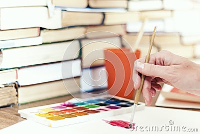 Female hand draws with a brush and watercolor paint on a sheet of paper on wooden table with stack of books background Stock Photo