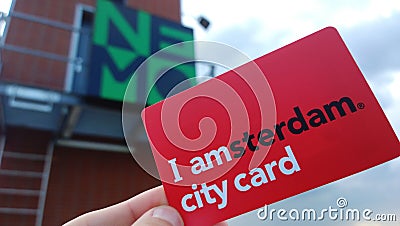 Female hand holding a red tourist guest card `I Amsterdam` on the background of signs of the Nemo Museum. Card for museums Editorial Stock Photo