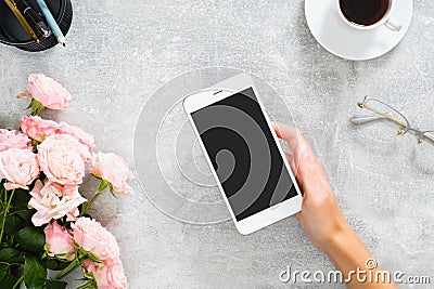 Female hand holding smartphone with blank screen mockup. Composition with roses flowers, coffee cup, stationery, glasses on Stock Photo