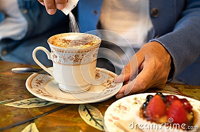 Female hand pouring sugar into cup of cappuccino in artistic ceramic mug. Breakfast with a small fresh fruit cake Stock Photo