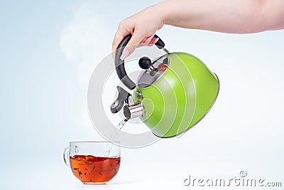 Female hand pours hot water from a green kettle into a transparent mug with a drink, on light blue background Stock Photo