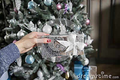 The female hand with red manicure holds a silvery box with a gift and a beautiful bow against the background of the New Year tree Stock Photo