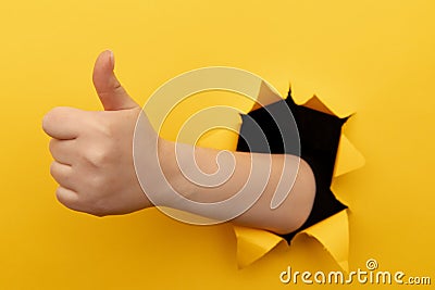 Female hand showing a thumb up sign through a ripped hole in yellow paper wall. Well done, good job concept Stock Photo