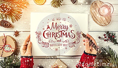 Female hand writing a letter to Santa. Christmas fir branches with holiday book, pencil, pine cones, gifts on white background. Stock Photo