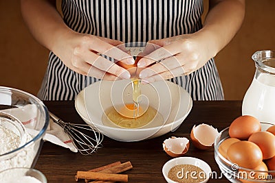 Female hands breaking egg into ceramic bowl on kitchen table with tableware. Stock Photo