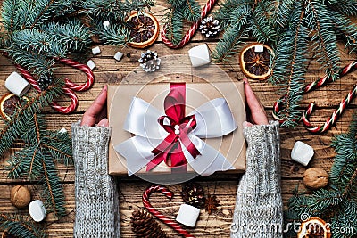 Female hands hold Gift box with bow. Christmas tree and decorations, cane caramel and oranges nuts Wooden background Stock Photo