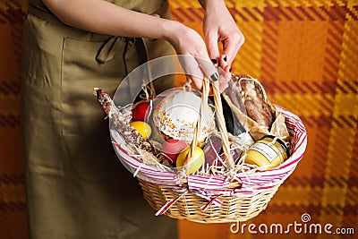 Female hands holding basket with colorful eggs, cake, red wine, hamon or jerky and dry smoked sausage. Food gift set for Stock Photo