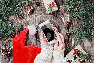 Female hands holding mobile phone on wooden background with Christmas gifts, Santa hat, Fir branches Stock Photo