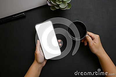 A female hands holding a smartphone mockup and a black coffee cup over modern black tabletop Stock Photo