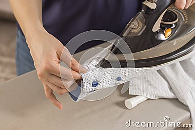 Female hands iron the shirt sleeve on a stand on an ironing board. Ironed clothes lie on the table in the background. Stock Photo