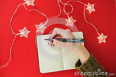 Female hands with manicure in green sweater write in notebook wish list, goals or promises for New Year on red Stock Photo