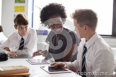Female High School Teacher Sitting At Table With Students Wearing Uniform Using Digital Tablets In Lesson Stock Photo