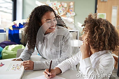 Female infant school teacher working one on one with a young schoolgirl, sitting at a table smiling at each other, front view, clo Stock Photo