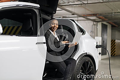 Female with laptop waiting inside EV at indoor charge point Stock Photo