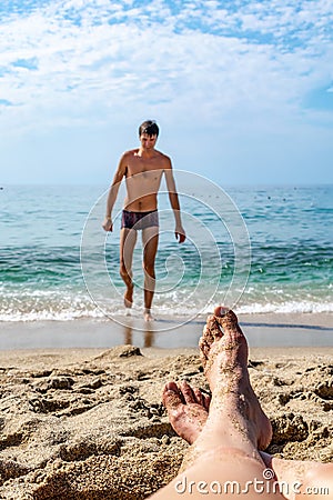 Female legs soiled with sand lie on the beach against the background of a blurred male silhouette emerging from the water. Couple Stock Photo