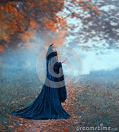 Female magician flees in the autumn orange forest, wearing a blue velvet cloak with a hood and wide sleeves. frozen Stock Photo